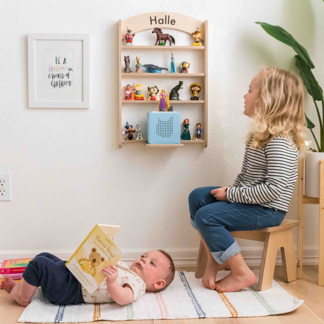 A tonie regal is mounted to a wall displaying its ability to hold and display the toniebox and figures that go with it. A baby is laying on the floor while a young girl is admiring her tonie shelf.