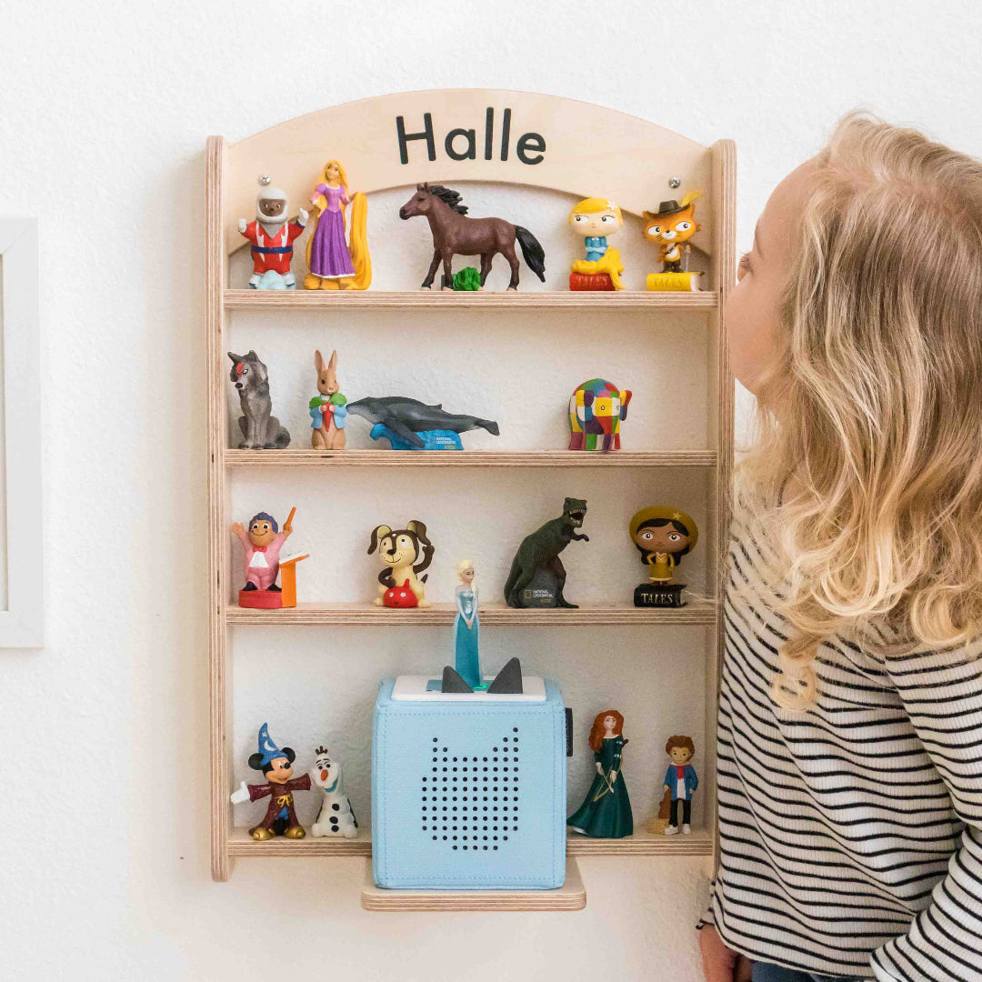 A young girl standing next to a tonie regal shelf that has her name lightly carved into the wood at the top of her shelf making it personal to her.