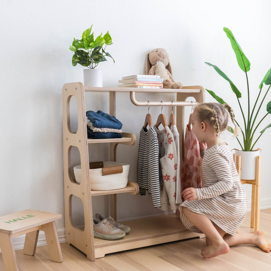 A little girl choosing clothes from a wooden toddler wardrobe with a curved hanging rod