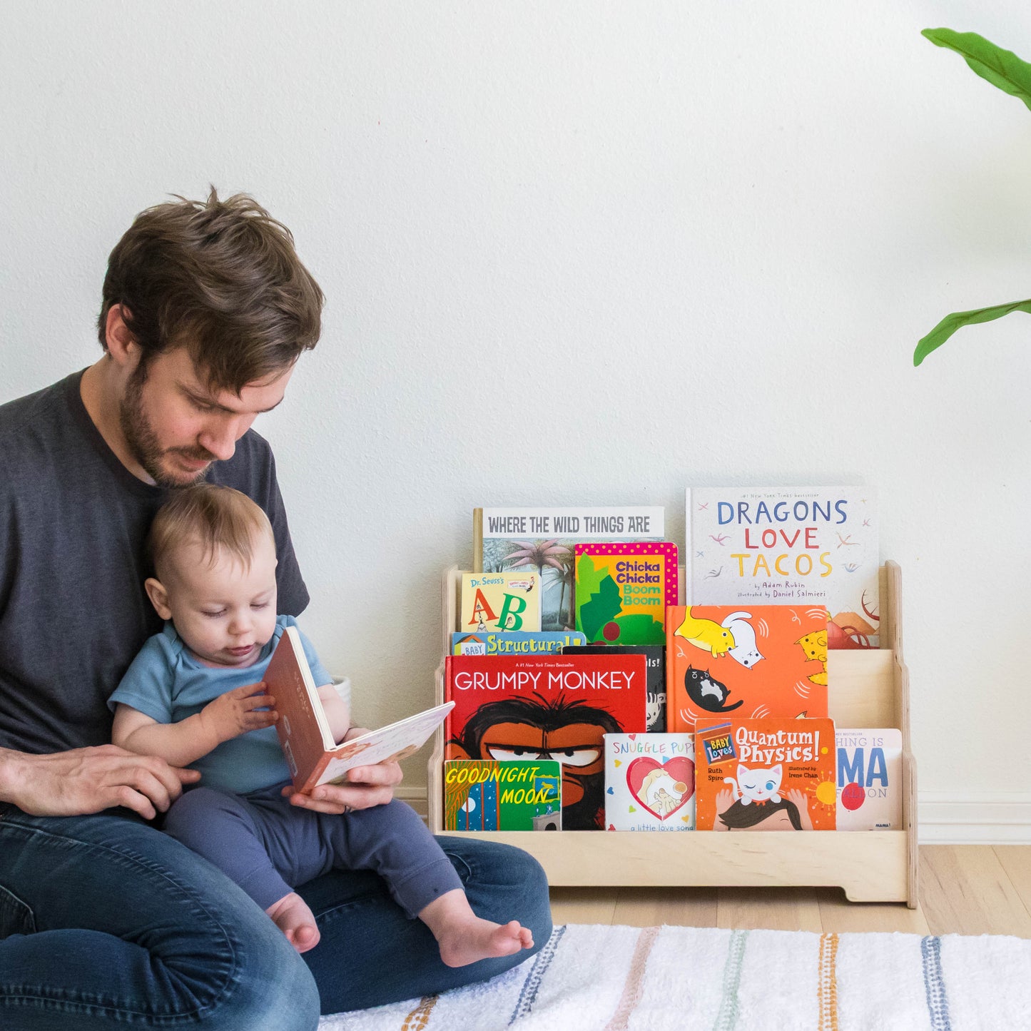 A father and child reading books next t a wooden bookshelf.    