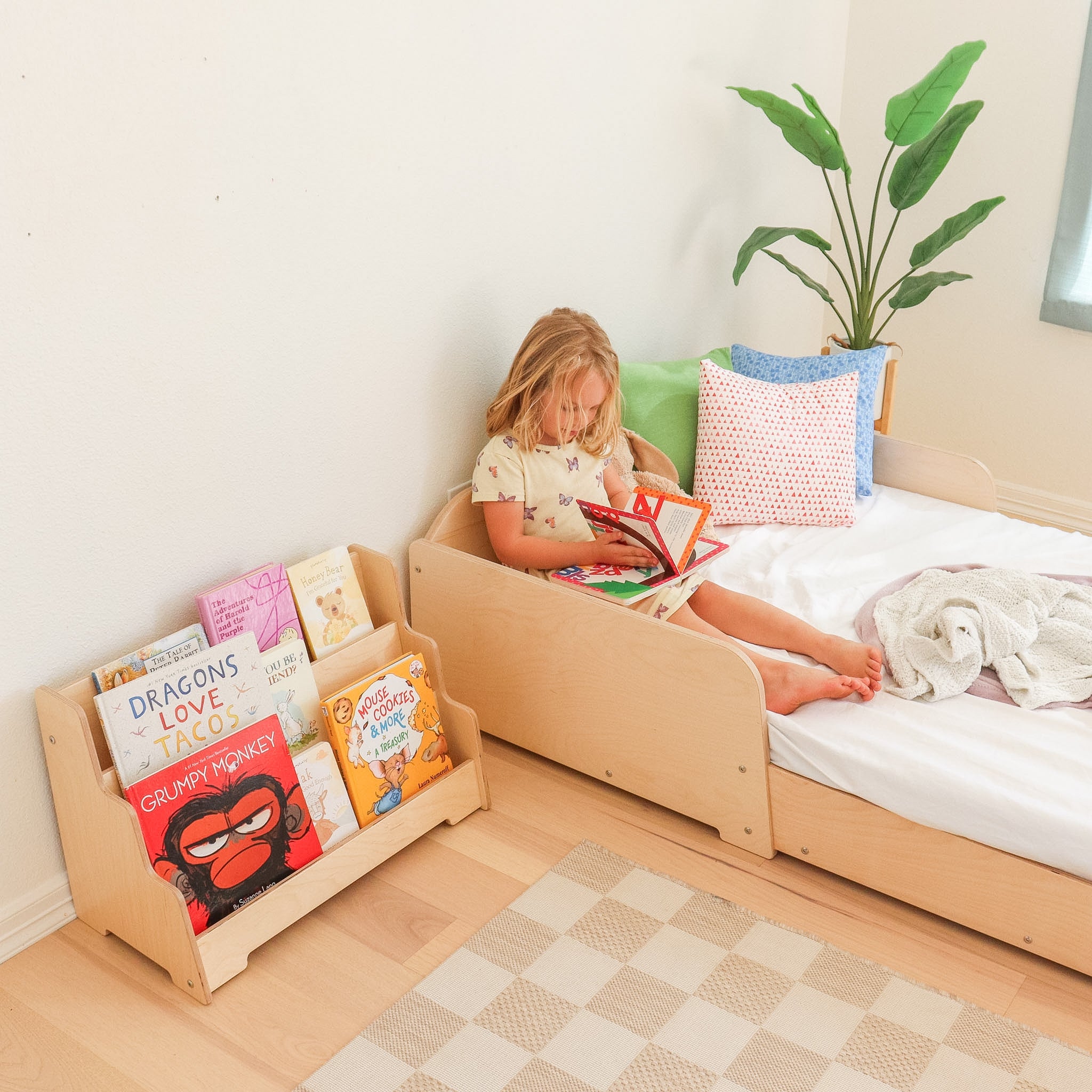 Girl reading a book while sitting on a kids wooden floor bed with a montessori bookshelf next o the floor bed. 