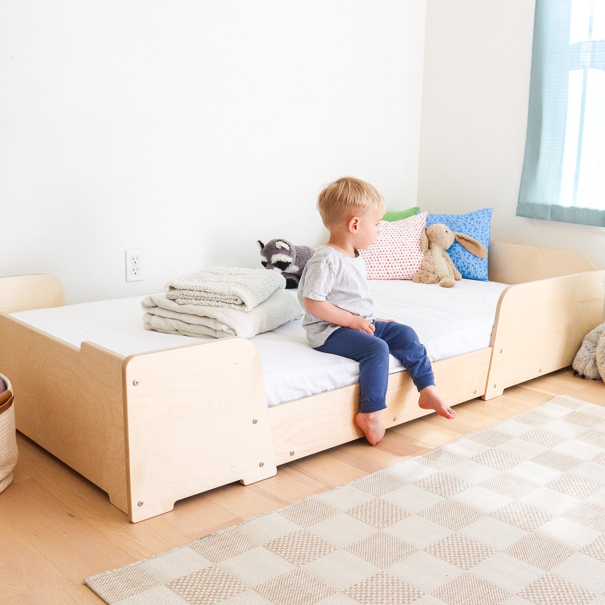 Child sitting on a wooden floor bed in a bright room with toys and pillows.