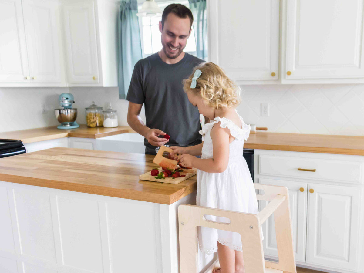 A dad and his daughter cutting strawberries together while she stands in a wooden learning tower at the kitchen counter
