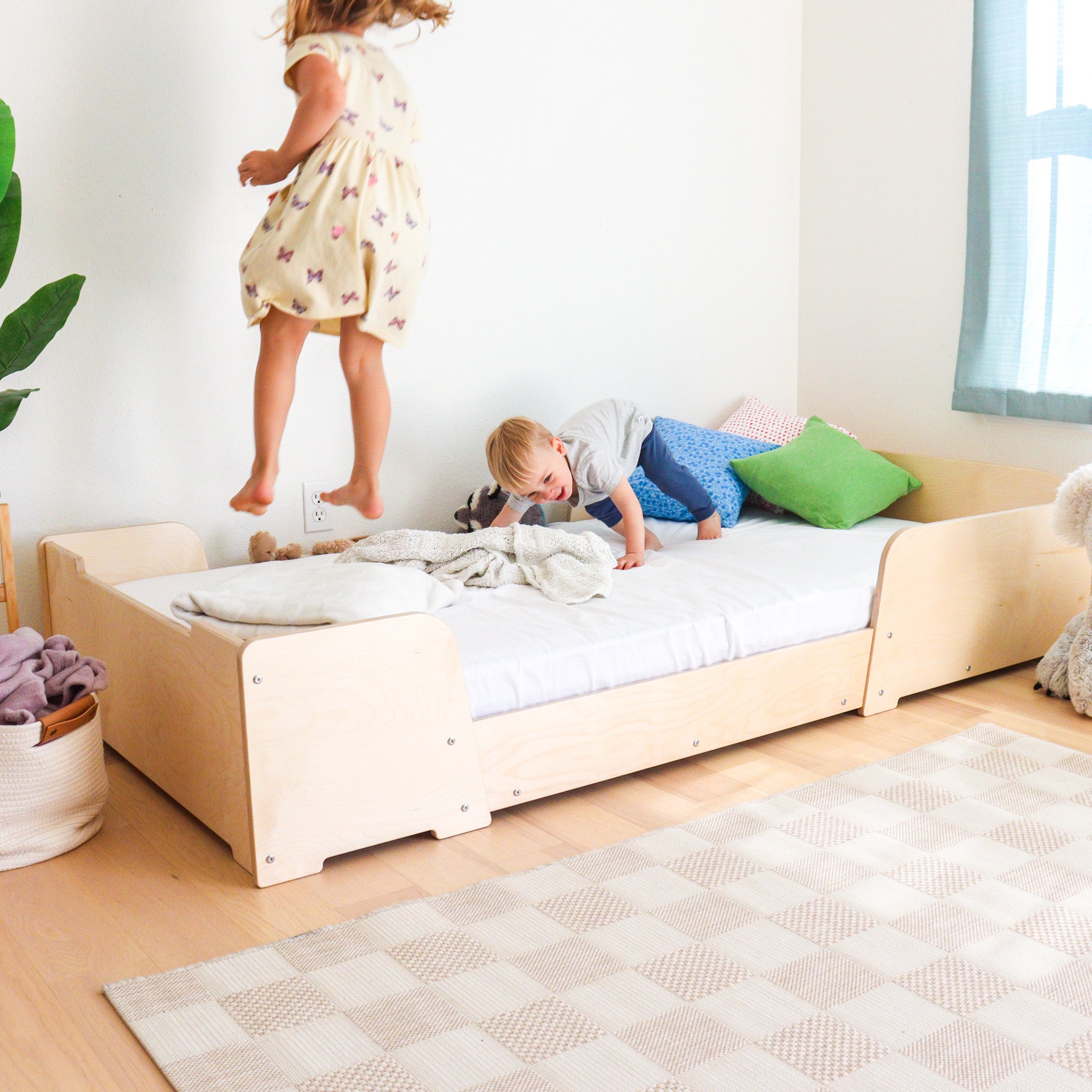 Children playing on a Montessori floor bed in a bright room with a plant and rug.