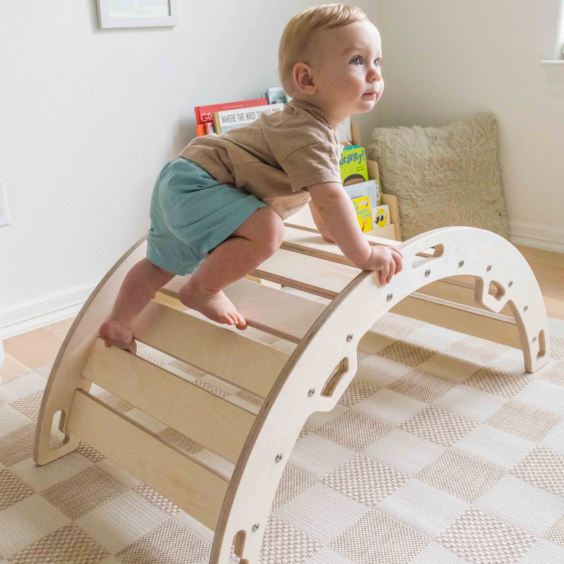Baby standing on a wooden climbing toy in a room.